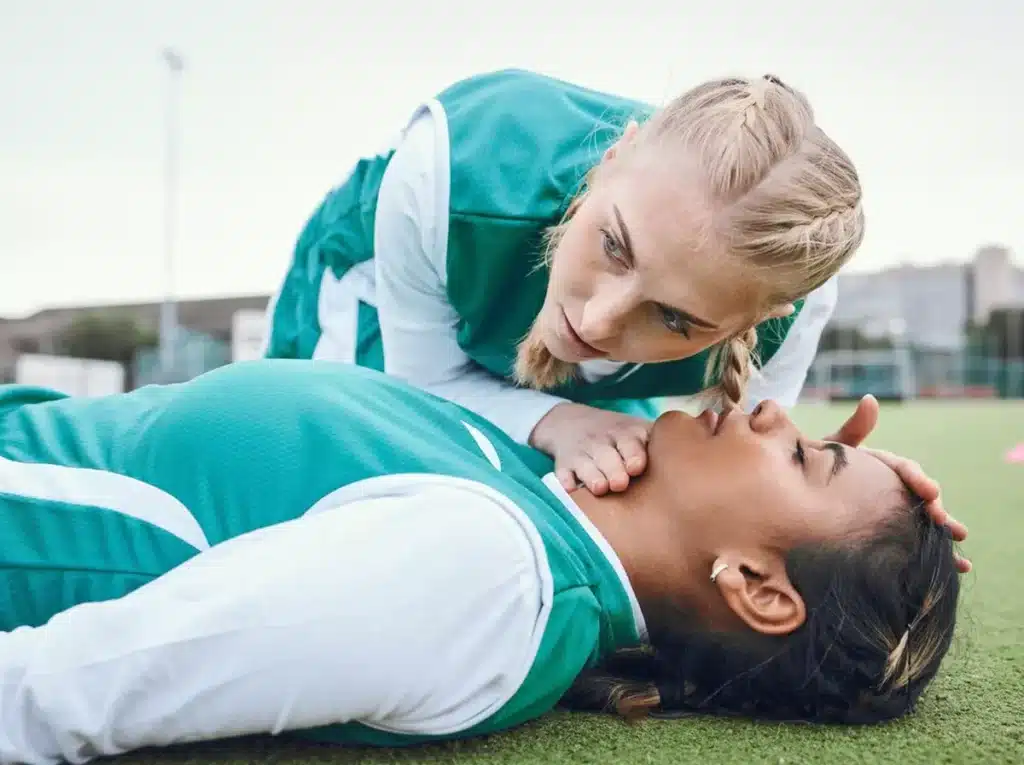 A female soccer player performing CPR on another soccer player, on a soccer field.