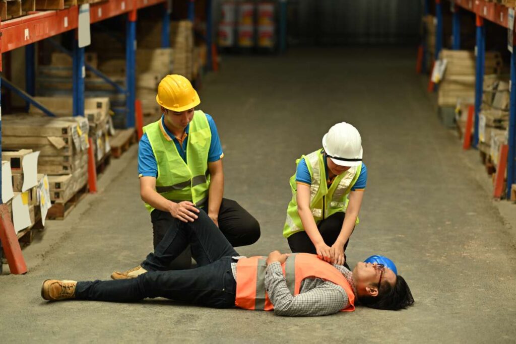 Warehouse workers kneeling next to a fallen man, preparing to perform CPR.